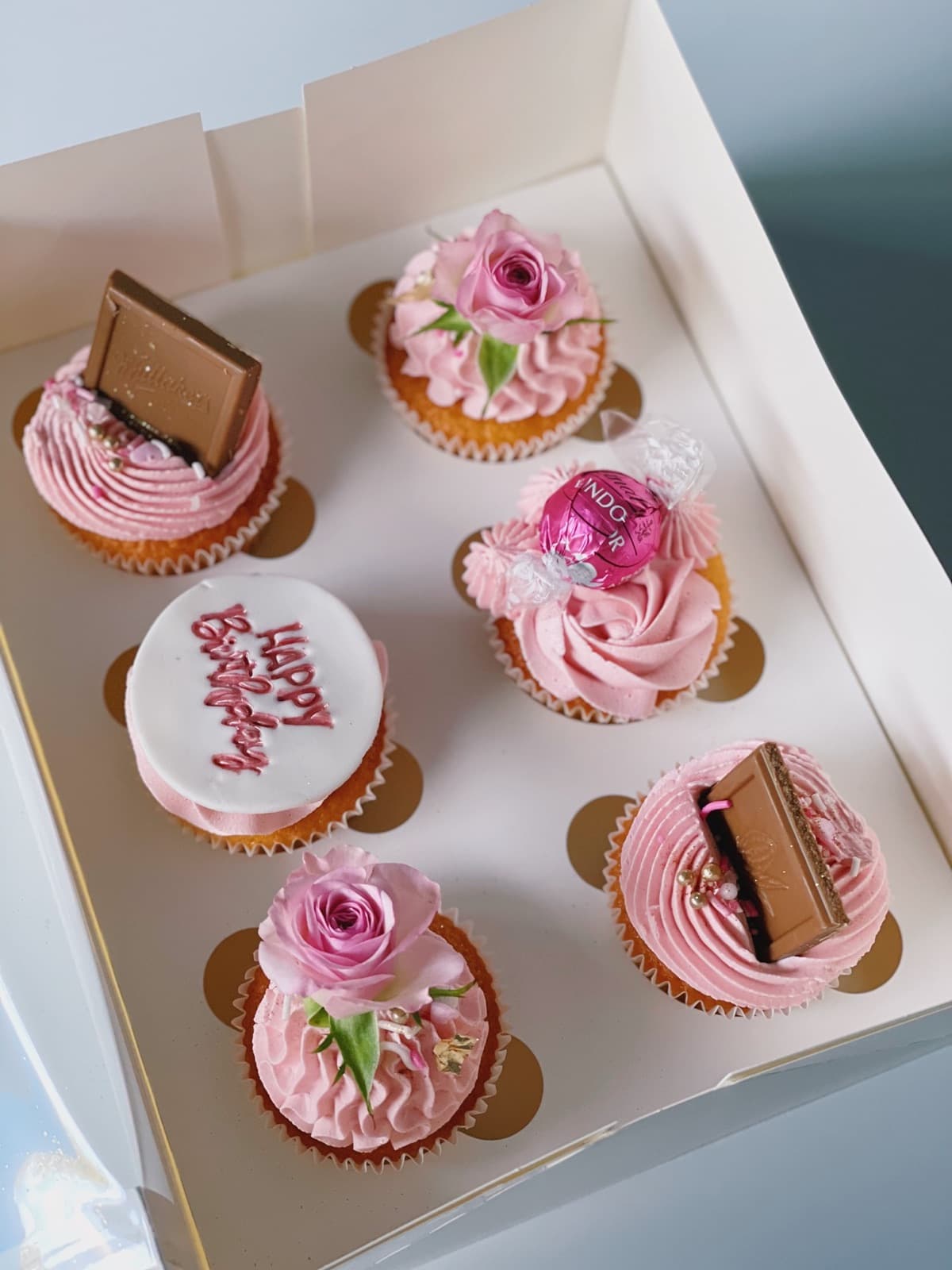 A boxed set of pink birthday cupcakes, each topped with a piped rosette.