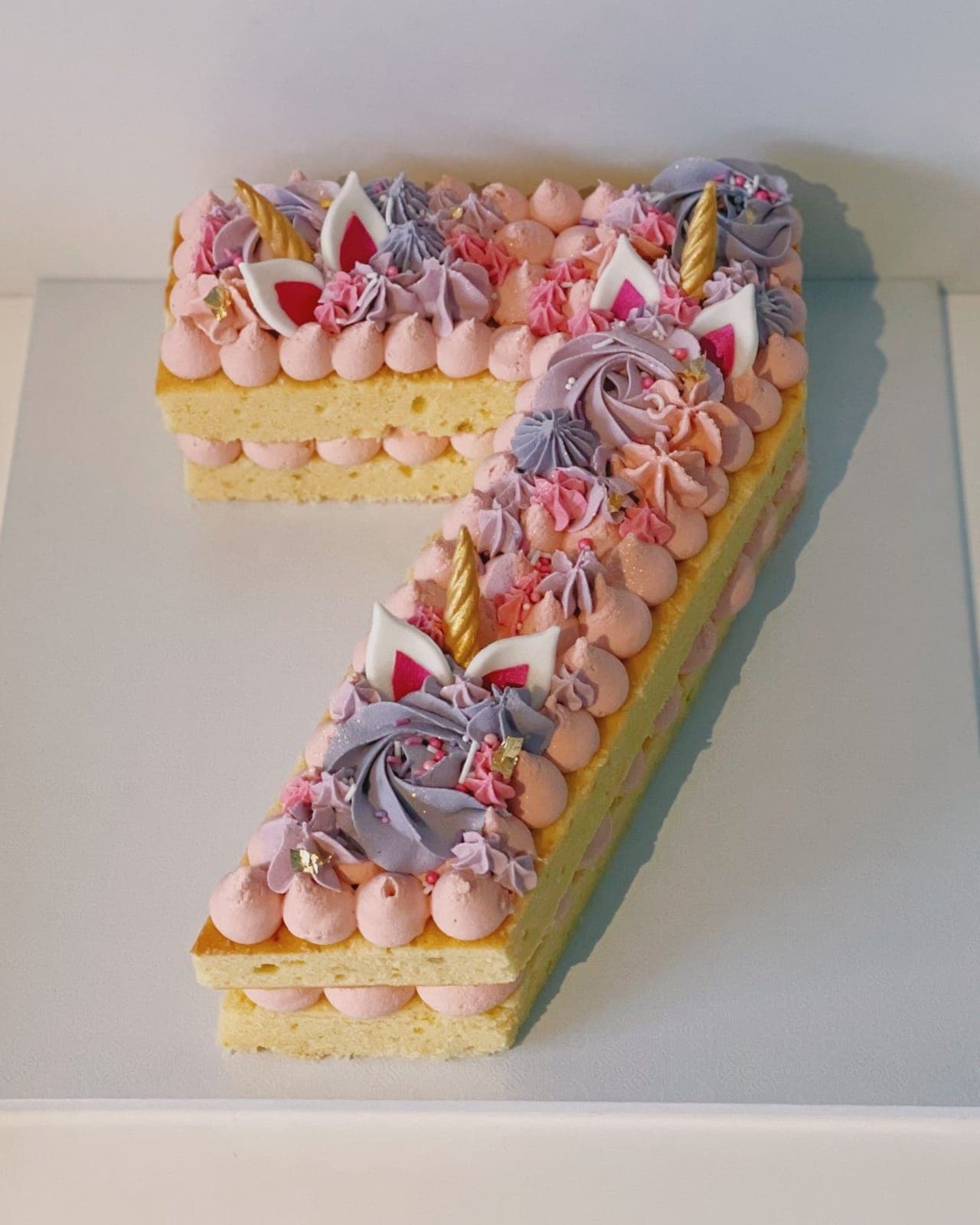 A cake cut in the shape of the number seven, two layers of sponge piped with pink and lilac kisses, topped with gold unicorn horns and ears.
