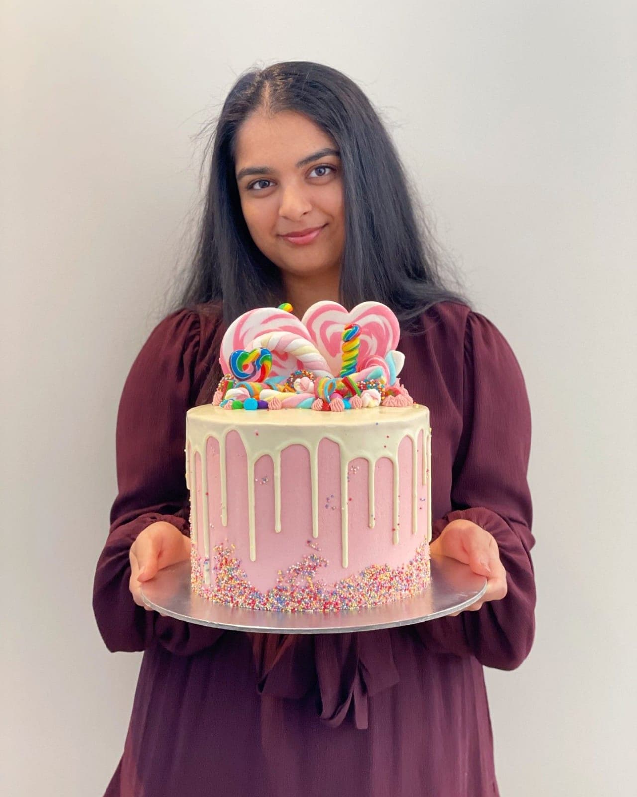 A pink drip cake held up for the camera, piled with swirl lollipops, marshmallow twists and lollies, with rainbow sprinkles around the base.