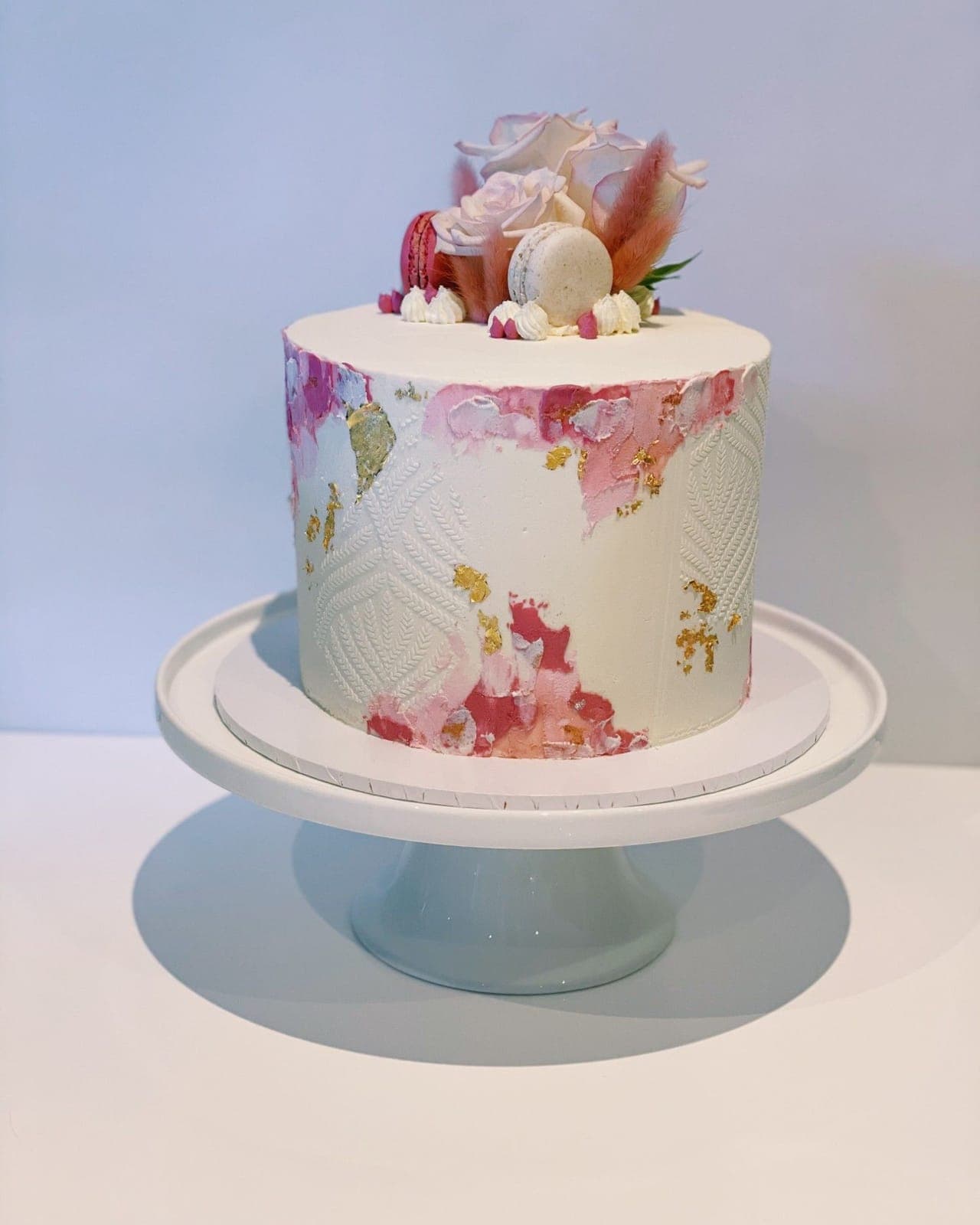A white cake with a stencilled leaf pattern, strokes of pink and flakes of gold leaf, topped with pale roses, macarons and dried grasses.