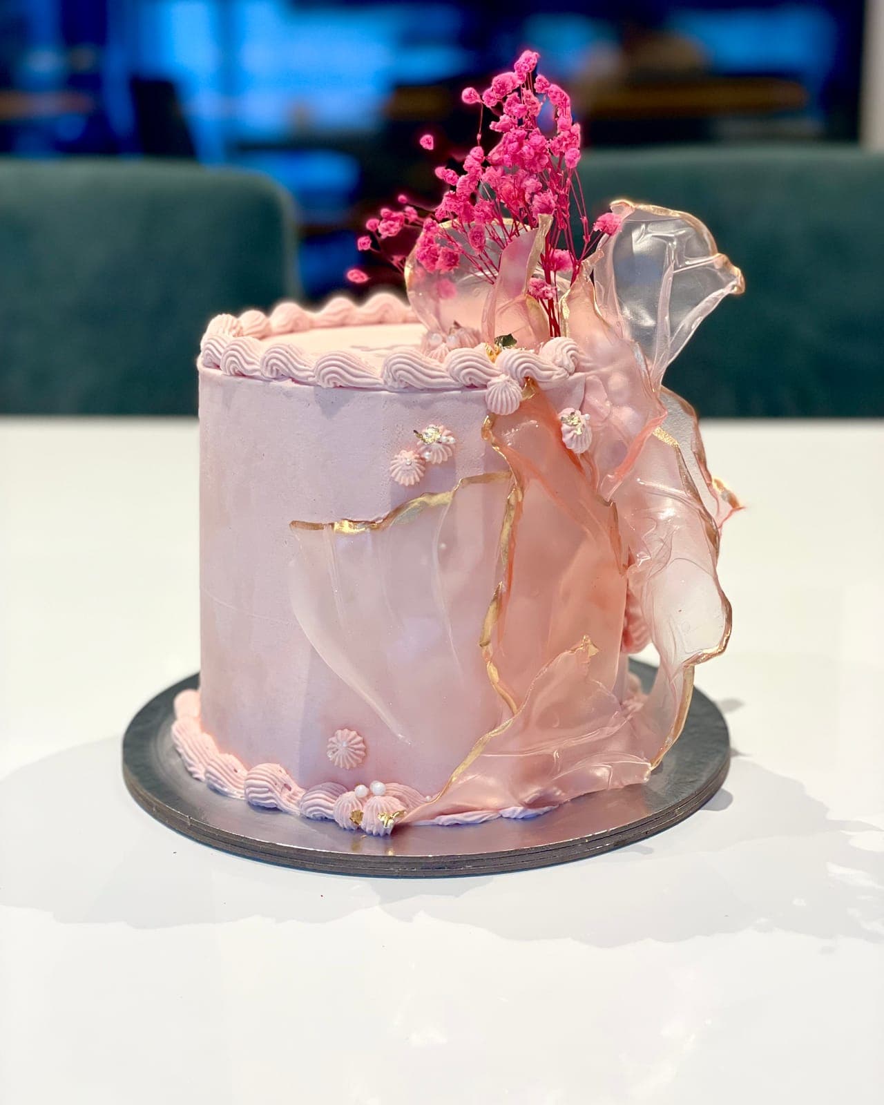 A tall blush-pink cake with fine gold lines, a sheer pink ruffle wrapped around one side and a spray of pink flowers on top.