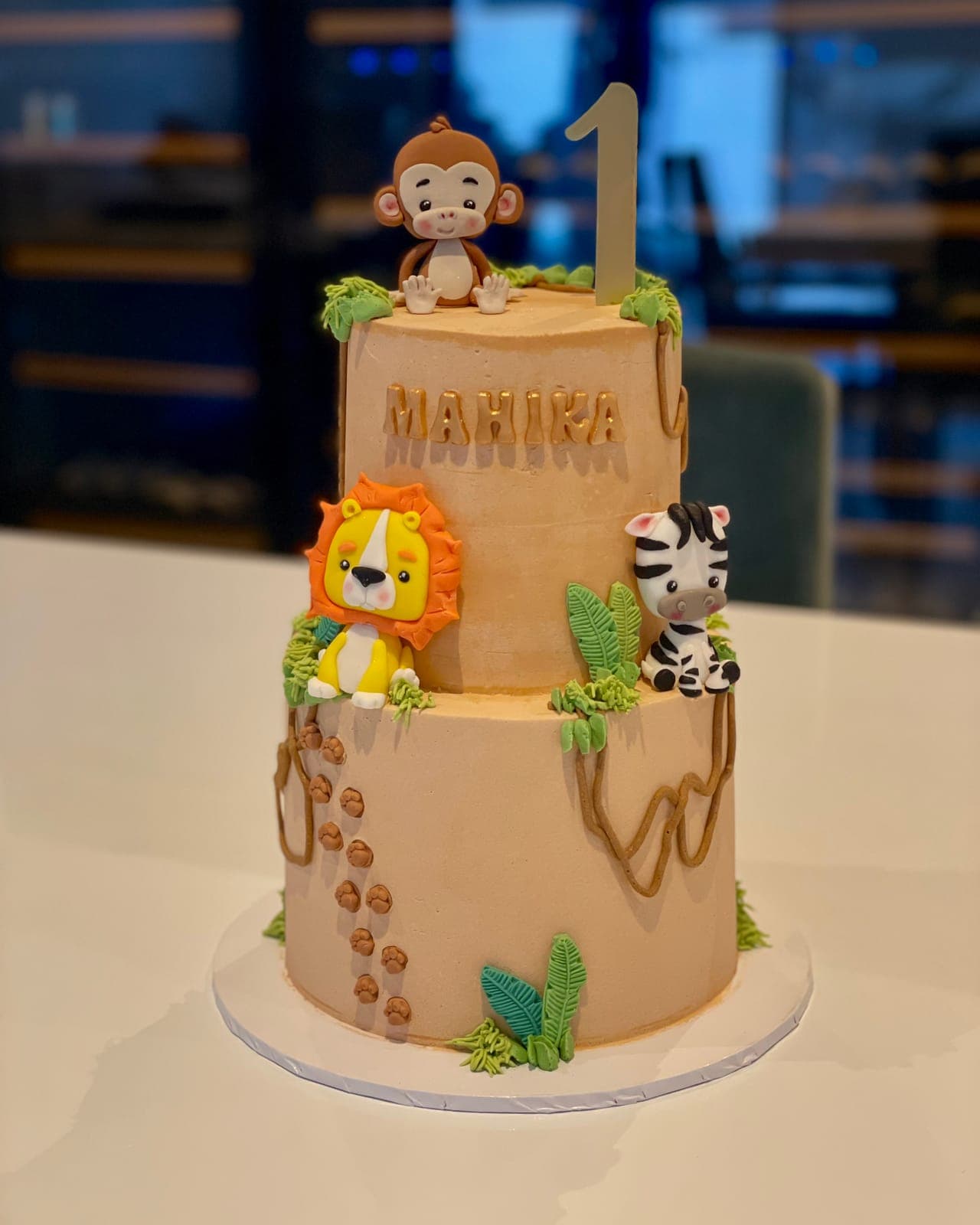A two-tier first-birthday cake in sandy tones with a modelled monkey, lion and zebra, jungle leaves and vines, and a gold number one topper.