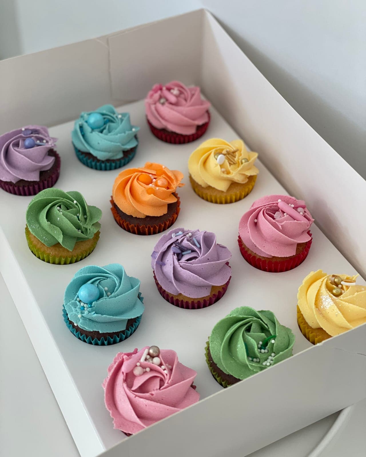 A box of cupcakes piped with rosettes in pastel purple, blue, green, orange, pink and yellow, finished with pearl sprinkles.