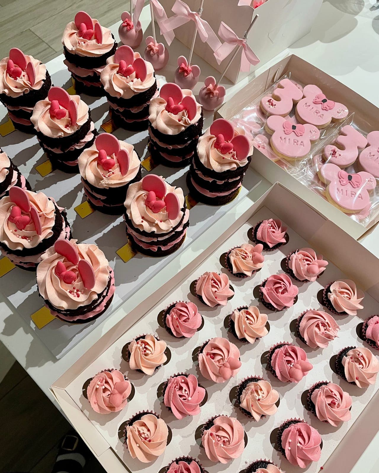Chocolate cupcakes piped with pink buttercream and topped with red bows, beside trays of pink rosette cupcakes and boxed pink cookies.