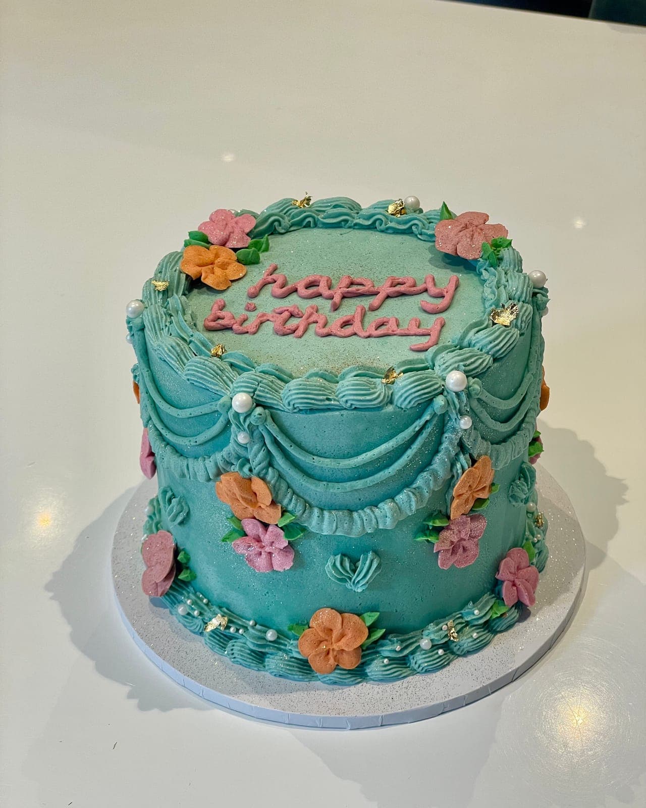 A turquoise cake with piped vintage swags, peach and pink piped flowers, and happy birthday written in pink script on top.