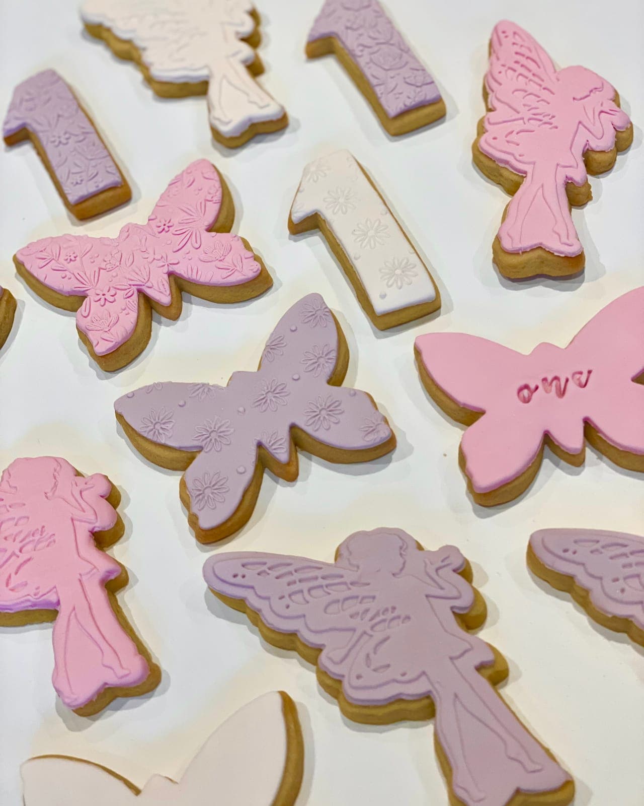 Cookies cut as fairies, butterflies and number ones, iced in pink, lilac and white with embossed patterns.